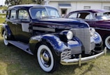 A vintage black Chevrolet car is parked on grass, with a shiny chrome grille and round headlights. The car has white-wall tires and a 1939 license plate. Another vintage car, maroon in color, is parked beside it.