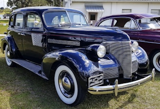 A vintage black Chevrolet car is parked on grass, with a shiny chrome grille and round headlights. The car has white-wall tires and a 1939 license plate. Another vintage car, maroon in color, is parked beside it.