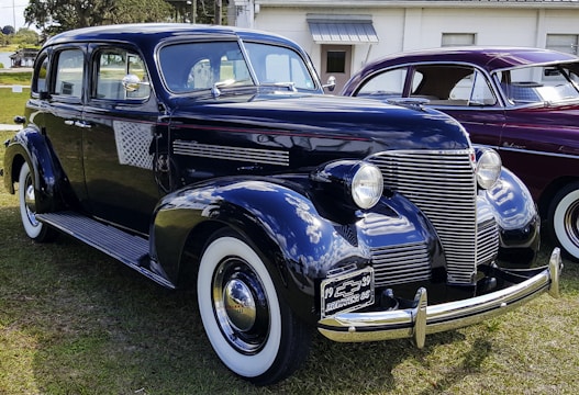 A vintage black Chevrolet car is parked on grass, with a shiny chrome grille and round headlights. The car has white-wall tires and a 1939 license plate. Another vintage car, maroon in color, is parked beside it.