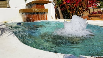 A small fountain with clear blue water creating splashes in a shallow rectangular pool. The fountain's cascade is lined with orange-brown bricks. Lush green and red tropical plants surround the fountain, with one large ceramic pot visible in the background. Bright sunlight casts shadows across the concrete.