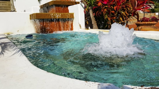 A small fountain with clear blue water creating splashes in a shallow rectangular pool. The fountain's cascade is lined with orange-brown bricks. Lush green and red tropical plants surround the fountain, with one large ceramic pot visible in the background. Bright sunlight casts shadows across the concrete.
