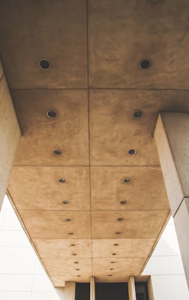 The image features a view of a structured ceiling composed of large, square beige panels with embedded round lights. The architectural design creates a geometric and uniform pattern, contributing to a modern, minimalist aesthetic.