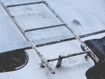 An old, rusted metal ladder is placed horizontally on a rough, cracked concrete surface with patches of black paint.