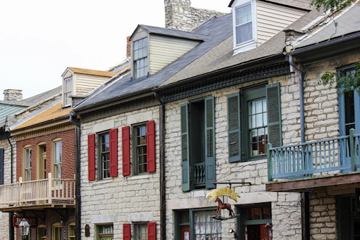 A row of historic buildings made from brick and stone, featuring shuttered windows and small porches. The architecture includes gabled roofs and dormer windows, with colors ranging from earthy tones to vibrant red shutters. The buildings have a quaint and rustic charm, typical of a small town or historic district.