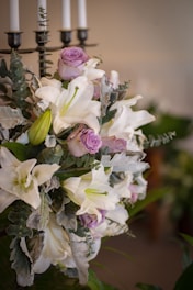 A floral arrangement featuring white lilies, light purple roses, and greenery. The bouquet is placed near a black candelabrum with white candles.