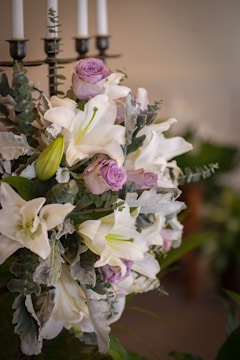 A floral arrangement featuring white lilies, light purple roses, and greenery. The bouquet is placed near a black candelabrum with white candles.