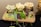 A wooden board holds three glasses filled with assorted cheese cubes, arranged alongside red grapes and garnished with fresh greens and slices of cucumber. The setup sits on a metal stand over a dark tablecloth, creating an elegant presentation of the food items.