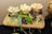 A wooden board holds three glasses filled with assorted cheese cubes, arranged alongside red grapes and garnished with fresh greens and slices of cucumber. The setup sits on a metal stand over a dark tablecloth, creating an elegant presentation of the food items.