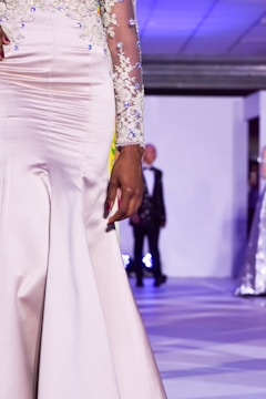 A close-up view of a person wearing an elegant cream-colored gown with intricate lace and sequin details on the sleeves. The background features a runway environment with another figure blurred in the distance.