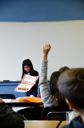 In a classroom setting, a student is standing at the front holding a large poster with illustrations and text. In the foreground, another student is seated at a desk with their hand raised. The classroom has a whiteboard and desks, and the overall environment is one of active learning.