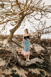A woman stands on a twisted and leafless tree branch, wearing sunglasses and a colorful sarong in a natural, beach-like setting. The scene conveys a sense of calm and connection with nature.
