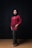 A person wearing a red traditional outfit with a patterned long skirt and a black hijab stands against a dark backdrop. The attire is accessorized with a statement necklace and bracelets.
