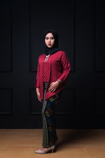 A person wearing a red traditional outfit with a patterned long skirt and a black hijab stands against a dark backdrop. The attire is accessorized with a statement necklace and bracelets.