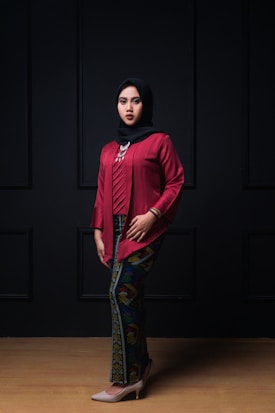 A person wearing a red traditional outfit with a patterned long skirt and a black hijab stands against a dark backdrop. The attire is accessorized with a statement necklace and bracelets.