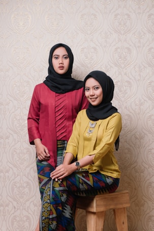 Two women wearing traditional attire with vibrant colors and black headscarves stand against a patterned wallpaper. One woman is seated on a wooden stool, while the other stands beside her.