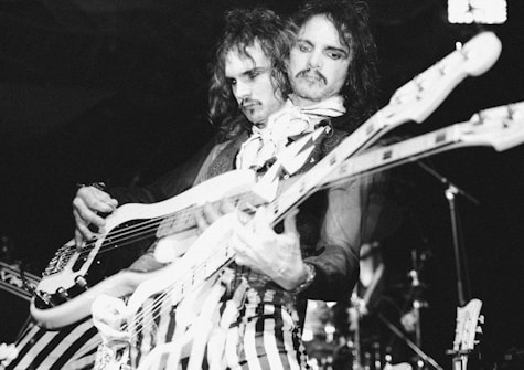 A musician playing an electric guitar with a psychedelic visual effect capturing two overlapping images of the person. The artist wears striped pants and a scarf, with a focused expression, suggesting intense concentration during the performance. The background appears to be dimly lit, hinting at a live concert setting.