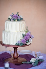 A two-tiered wedding cake with a smooth white frosting is elegantly adorned with purple and white roses, as well as greenery. The cake is placed on a round wooden cake stand and surrounded by a purple cloth. Small candles are positioned around the base along with additional floral arrangements.