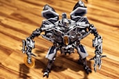A detailed robotic figure stands on a wooden surface. The robot features intricate metallic components, including articulated arms and legs, and a complex head design with visible mechanics. Its overall aesthetic is metallic, with a prominent gray and silver color scheme accented with darker and lighter shades. The robot appears poised and ready for action, with one arm equipped with a circular saw-like mechanism.