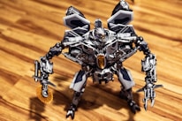 A detailed robotic figure stands on a wooden surface. The robot features intricate metallic components, including articulated arms and legs, and a complex head design with visible mechanics. Its overall aesthetic is metallic, with a prominent gray and silver color scheme accented with darker and lighter shades. The robot appears poised and ready for action, with one arm equipped with a circular saw-like mechanism.