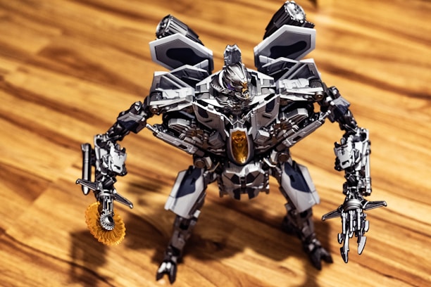 A detailed robotic figure stands on a wooden surface. The robot features intricate metallic components, including articulated arms and legs, and a complex head design with visible mechanics. Its overall aesthetic is metallic, with a prominent gray and silver color scheme accented with darker and lighter shades. The robot appears poised and ready for action, with one arm equipped with a circular saw-like mechanism.