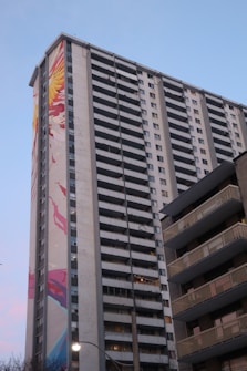 A tall residential building with multiple balconies, featuring a large, colorful mural on its side. The mural includes bright abstract shapes in red, yellow, and blue tones. The sky in the background is clear with a light blue hue.
