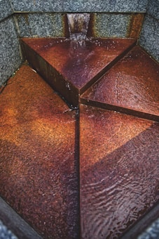 A triangular-shaped stone fountain with water flowing from a narrow spout at the top. The interior of the fountain is divided into three sections, with water trickling down the rusty, textured surface. The fountain is surrounded by gray stone walls.
