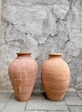 Two large, rustic clay pots with smooth, rounded surfaces are positioned side by side on a tiled ground. The backdrop is a weathered, textured concrete wall with visible cracks and uneven surfaces, adding a sense of age and authenticity to the scene.