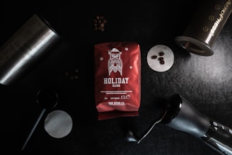 A red bag of Holiday Blend coffee is surrounded by various coffee-making tools on a dark surface. The bag features an illustration of an owl and snowflakes. Scattered coffee beans and an Aeropress, a cylindrical coffee grinder, and a filter paper add to the coffee-making theme.