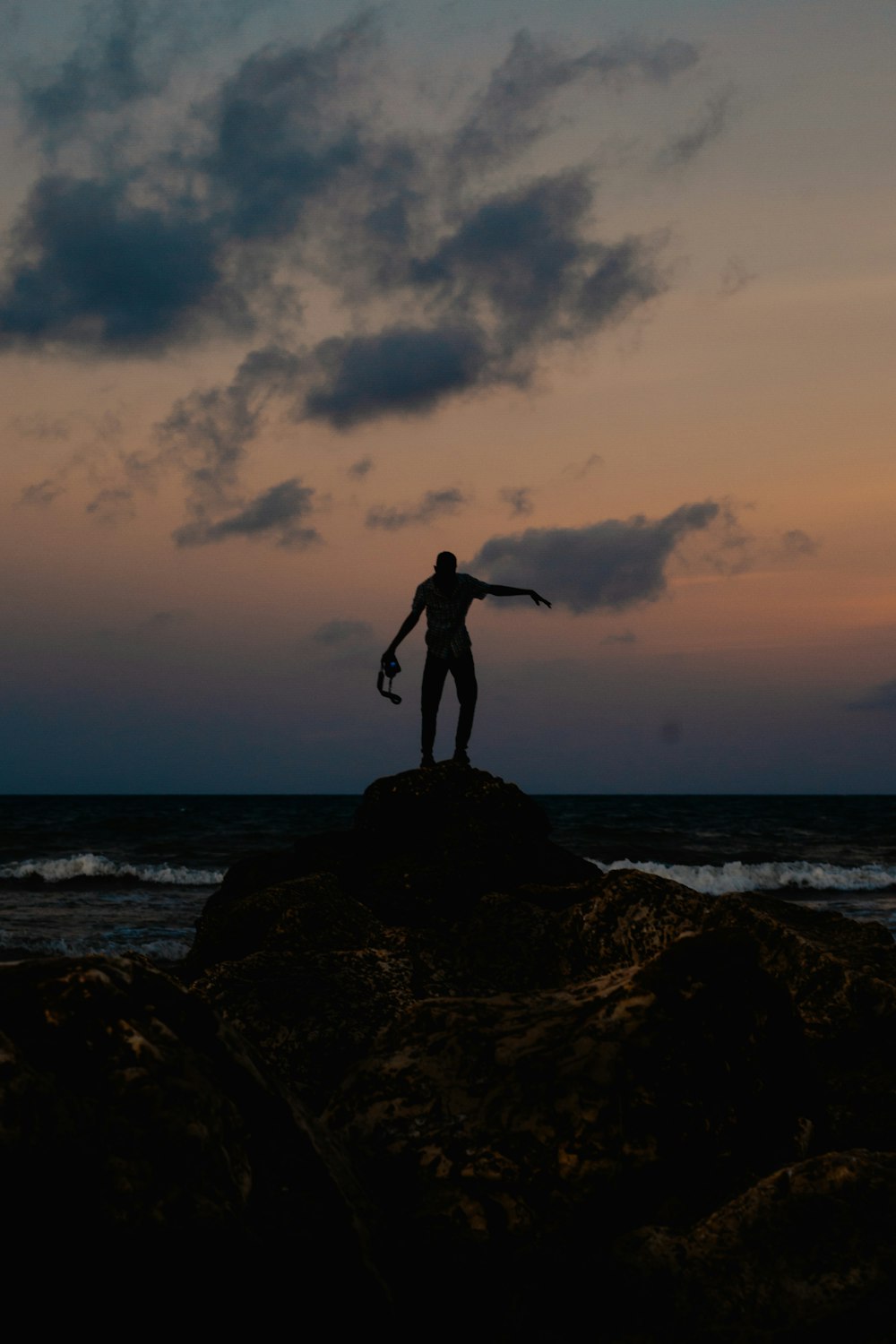 silhouette of man standing on rock formation near body of water during sunset