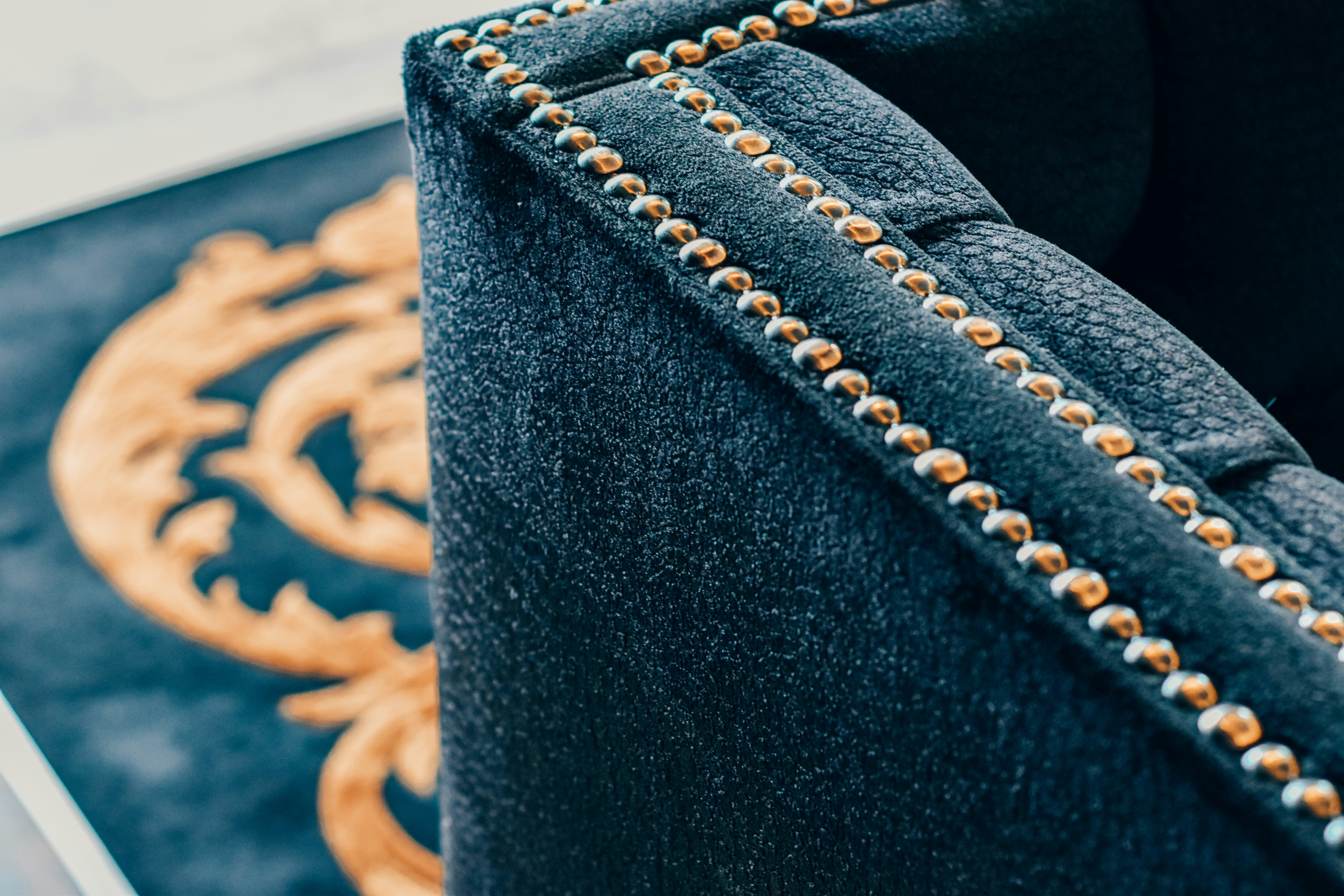 A close-up of a luxurious dark fabric upholstered piece of furniture with brass or golden studs aligned along the edges. In the background, part of a decorative pattern in gold on a black surface is visible.