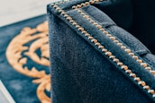 A close-up of a luxurious dark fabric upholstered piece of furniture with brass or golden studs aligned along the edges. In the background, part of a decorative pattern in gold on a black surface is visible.