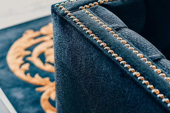 A close-up of a luxurious dark fabric upholstered piece of furniture with brass or golden studs aligned along the edges. In the background, part of a decorative pattern in gold on a black surface is visible.