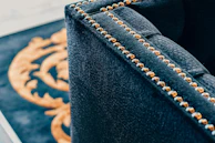 A close-up of a luxurious dark fabric upholstered piece of furniture with brass or golden studs aligned along the edges. In the background, part of a decorative pattern in gold on a black surface is visible.