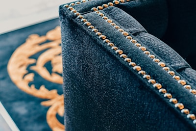 A close-up of a luxurious dark fabric upholstered piece of furniture with brass or golden studs aligned along the edges. In the background, part of a decorative pattern in gold on a black surface is visible.