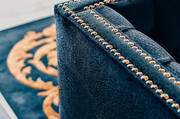 A close-up of a luxurious dark fabric upholstered piece of furniture with brass or golden studs aligned along the edges. In the background, part of a decorative pattern in gold on a black surface is visible.