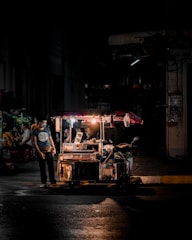 A street food vendor cart is illuminated by a single light in a dimly lit urban area. A person wearing a mask stands beside the cart, while a few others are dimly visible in the background. The setting appears to be a nighttime street scene.
