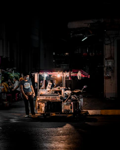 A masked volunteer in a black hoodie handing a warm meal to a person sitting on a curb under dim streetlights with subtle smoke in the background.