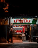 A small, warmly lit street-level shop with a prominent sign in a foreign script. The shop features bright red and green colors with a light hanging above the entrance. Inside, there are subtle hints of activity or items, such as hanging cloths or posters. The overall scene appears cozy and inviting, typical of an evening setting.