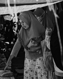 A black-and-white image of a market scene where women wearing hijabs are interacting with the items on display. One woman is pointing at something on a table while holding a small bag under her other arm. Another woman is standing next to her, and there are several other people in the background, partially visible.