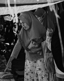A black-and-white image of a market scene where women wearing hijabs are interacting with the items on display. One woman is pointing at something on a table while holding a small bag under her other arm. Another woman is standing next to her, and there are several other people in the background, partially visible.