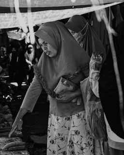 A black-and-white image of a market scene where women wearing hijabs are interacting with the items on display. One woman is pointing at something on a table while holding a small bag under her other arm. Another woman is standing next to her, and there are several other people in the background, partially visible.
