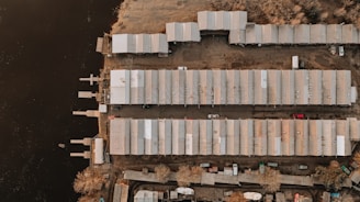 An aerial view of a waterfront area with rows of industrial or storage buildings placed parallel to a body of water. The buildings have metal roofs and are flanked by parking areas where several vehicles are visible. Sparse vegetation is present around the outer edges, and small docks extend into the water.