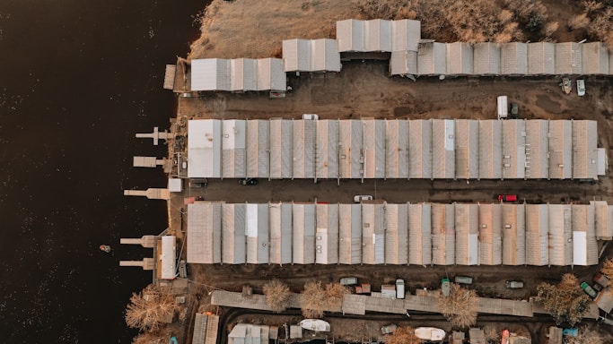 An aerial view of a waterfront area with rows of industrial or storage buildings placed parallel to a body of water. The buildings have metal roofs and are flanked by parking areas where several vehicles are visible. Sparse vegetation is present around the outer edges, and small docks extend into the water.