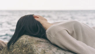 woman in white knit sweater lying on brown rock near sea during daytime