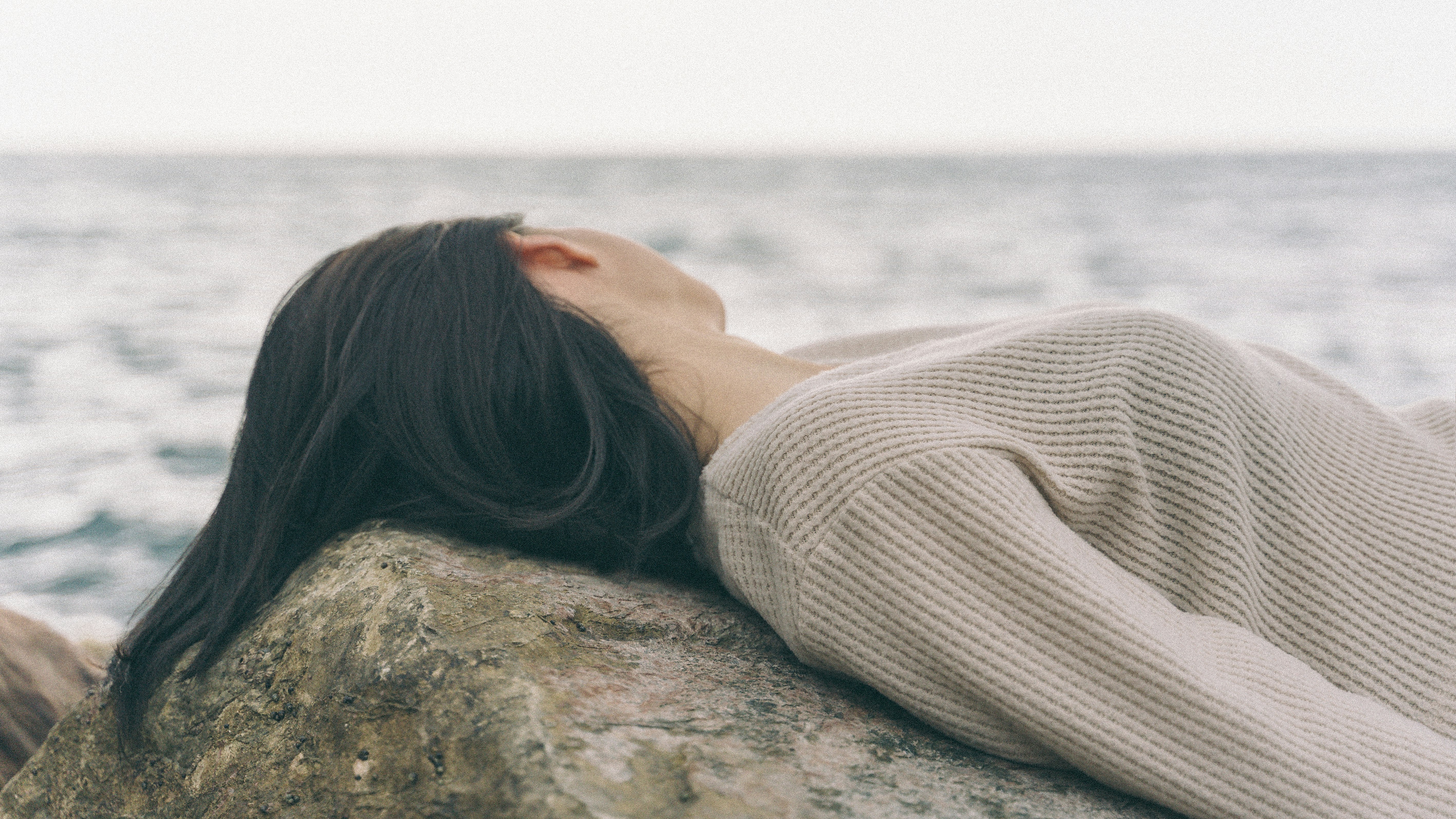 woman in white knit sweater lying on brown rock near sea during daytime