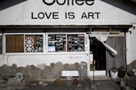 A storefront with a sign that reads 'LOVE IS ART' above the entrance. The facade features a stone wall with various decorations. The window displays several old-fashioned cameras and other vintage objects. A partially open wooden door with a curtain is on the right side. Various signs with text are placed outside, and there are a few pottery pieces near the entrance.