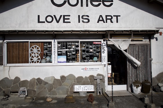 A storefront with a sign that reads 'LOVE IS ART' above the entrance. The facade features a stone wall with various decorations. The window displays several old-fashioned cameras and other vintage objects. A partially open wooden door with a curtain is on the right side. Various signs with text are placed outside, and there are a few pottery pieces near the entrance.