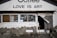 A storefront with a sign that reads 'LOVE IS ART' above the entrance. The facade features a stone wall with various decorations. The window displays several old-fashioned cameras and other vintage objects. A partially open wooden door with a curtain is on the right side. Various signs with text are placed outside, and there are a few pottery pieces near the entrance.