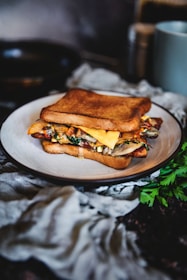 A delicious-looking sandwich with toasted bread, filled with melted cheese, vegetables, and possibly bacon or similar meat, sitting on a white plate. The setting includes a dark background with some green parsley and a soft cloth nearby, creating a rustic and cozy atmosphere.