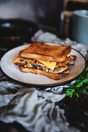 A delicious-looking sandwich with toasted bread, filled with melted cheese, vegetables, and possibly bacon or similar meat, sitting on a white plate. The setting includes a dark background with some green parsley and a soft cloth nearby, creating a rustic and cozy atmosphere.